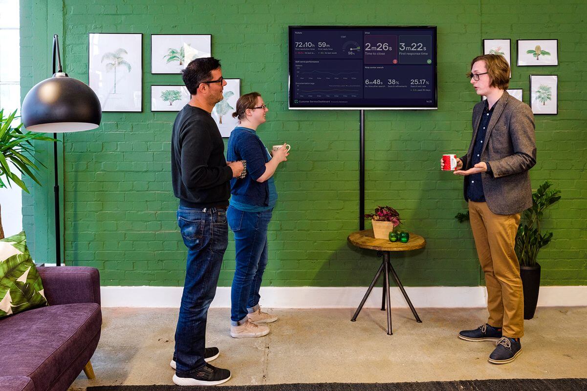 Three people standing in front of a wall-mounted TV dashboard